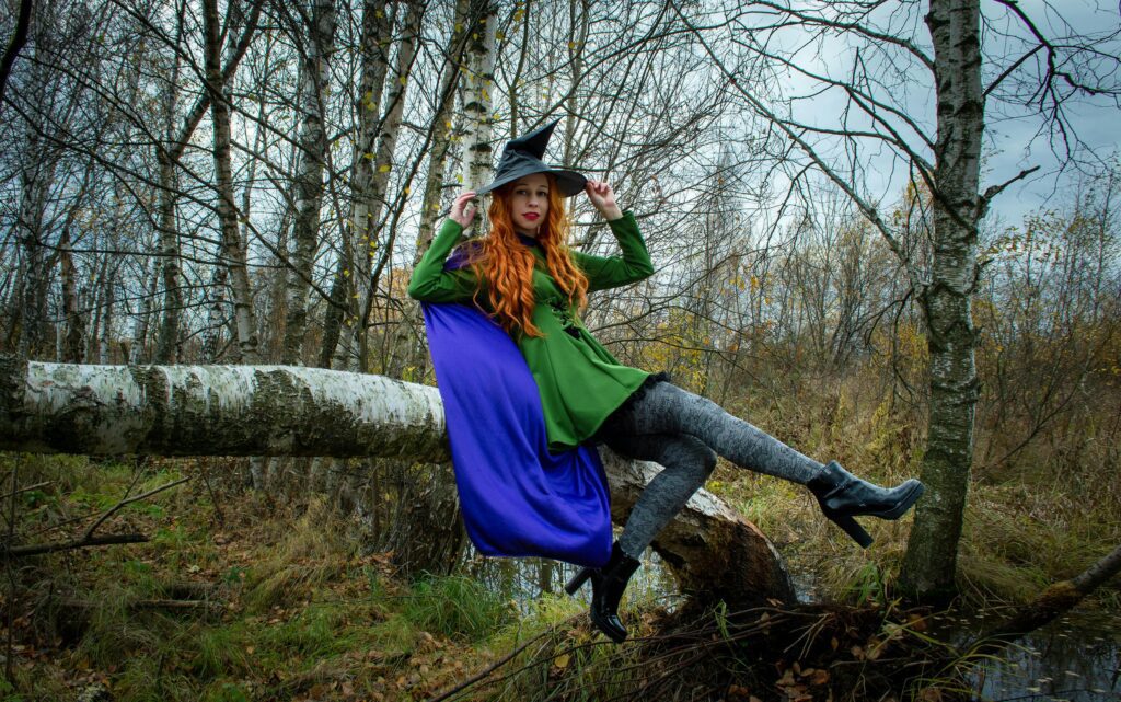 A woman in a witch costume poses on a tree in an autumn forest.