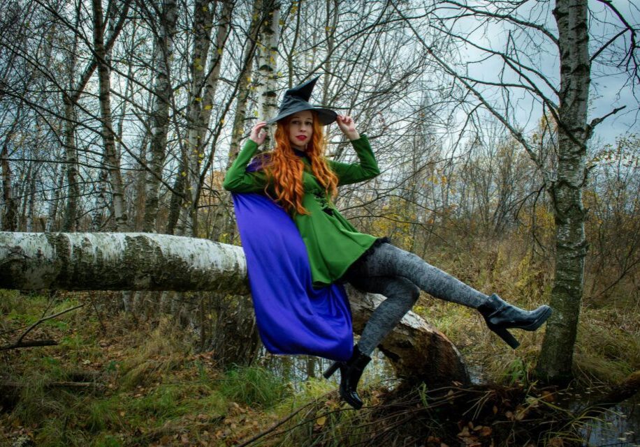 A woman in a witch costume poses on a tree in an autumn forest.